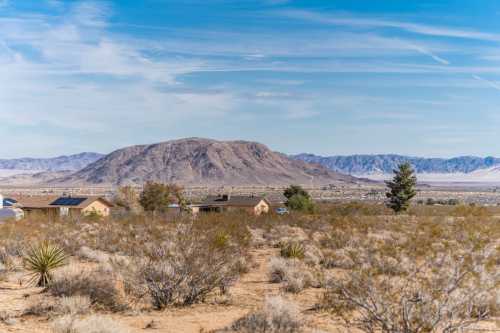 A desert landscape with sparse vegetation, houses, and a distant mountain under a blue sky with wispy clouds.