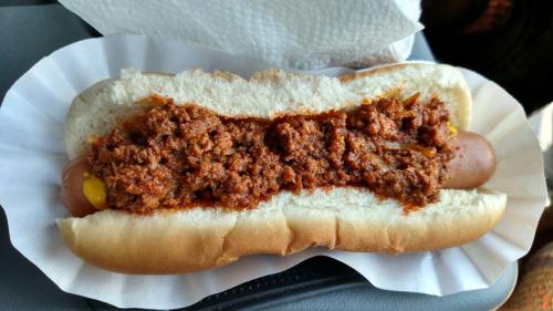 A hot dog in a bun topped with a generous serving of chili and mustard, served on a paper tray.