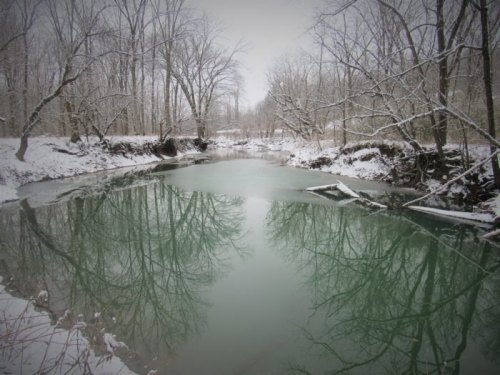 A serene winter scene of a river surrounded by snow-covered trees, with reflections on the water's surface.