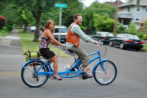 A man and woman joyfully ride a blue tandem bike on a suburban street, surrounded by trees and parked cars.