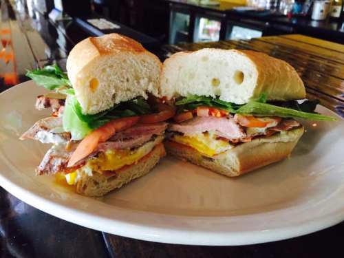 A freshly cut sandwich with layers of meat, lettuce, tomato, and egg on a crusty roll, served on a white plate.
