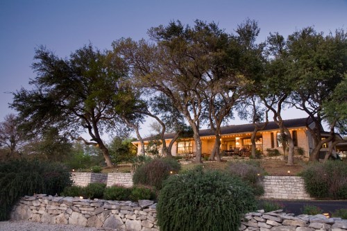 A cozy house nestled among trees, with warm lights glowing at dusk and a stone wall in the foreground.
