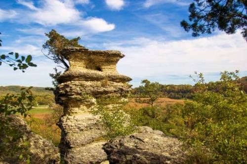 A unique rock formation stands tall amidst lush greenery and a clear blue sky, with rolling hills in the background.