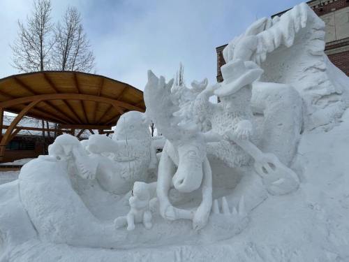 A detailed snow sculpture featuring a moose and other animals, set under a wooden pavilion against a cloudy sky.