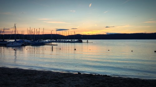 A serene sunset over a calm lake, with sailboats docked and a sandy beach in the foreground.