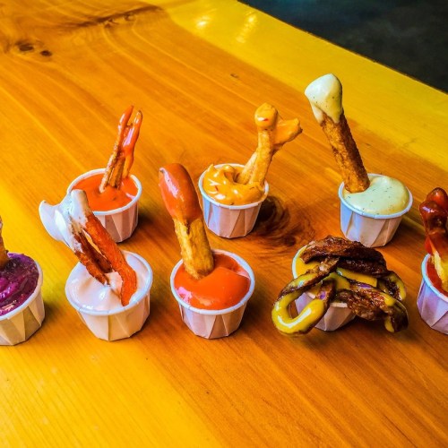 A variety of dipping sauces in small cups with pretzel sticks, arranged on a wooden table.