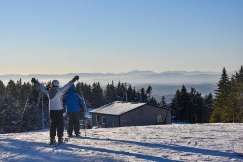 Two skiers with arms raised walk towards a ski lodge, surrounded by snow-covered trees and distant mountains.
