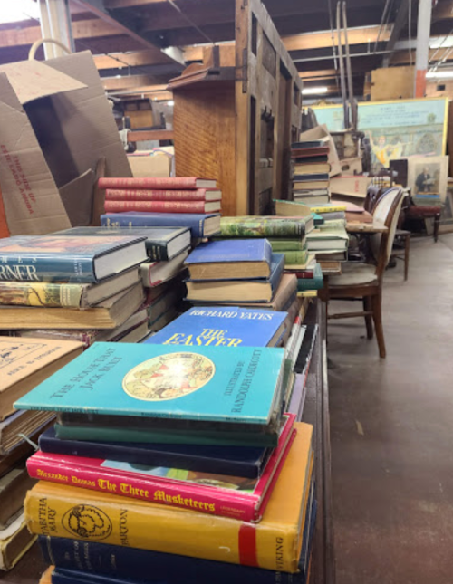 A cluttered table filled with colorful books in a vintage shop, with wooden furniture and boxes in the background.