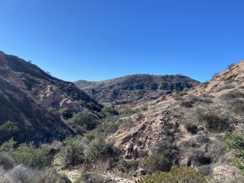 A scenic view of rolling hills and a dry valley under a clear blue sky, with sparse vegetation and rocky terrain.