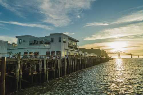 A waterfront view of a building on a pier at sunset, with a bridge visible in the background and calm water in the foreground.