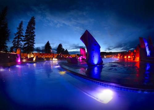 A serene outdoor pool at dusk, illuminated by colorful lights and surrounded by trees and stone sculptures.