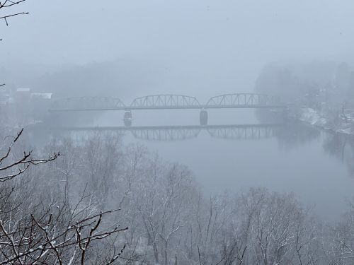 A foggy winter scene featuring a bridge over a river, surrounded by snow-covered trees and a misty atmosphere.