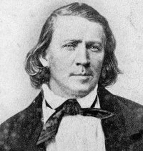 Black and white portrait of a man with long hair, wearing a suit and bow tie, looking directly at the camera.