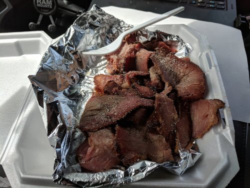A foil-wrapped container filled with sliced, seasoned smoked meat, resting on a white takeout tray.