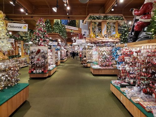 A festive store aisle filled with Christmas decorations, trees, and ornaments, with shoppers in the background.