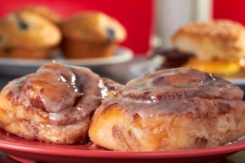 Two gooey cinnamon rolls with icing on a red plate, with muffins and a biscuit in the background.