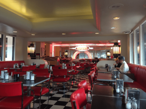 A retro diner interior with red booths, checkered floor, and neon lights, featuring a few patrons seated at tables.