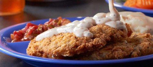 A blue plate with crispy fried chicken topped with creamy gravy, accompanied by salsa and a side of vegetables.