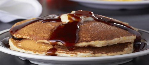 A stack of golden pancakes drizzled with syrup and topped with a pat of butter on a white plate.