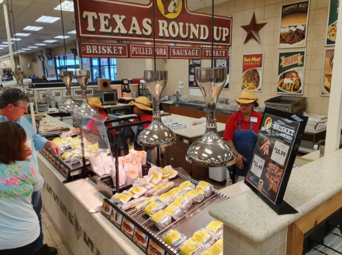 A busy deli counter with various meats and sides, featuring staff in hats and customers selecting food.