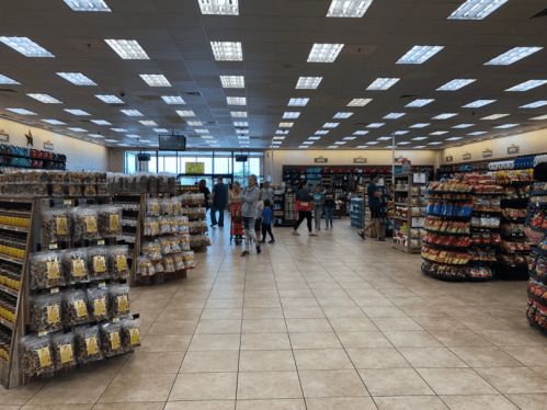 A spacious store interior with shelves of snacks and customers browsing in the aisles. Brightly lit with tile flooring.