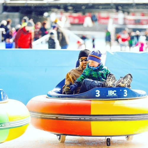 Two children enjoy tubing on a snowy slope, surrounded by other people in winter attire. Brightly colored tubes are visible.