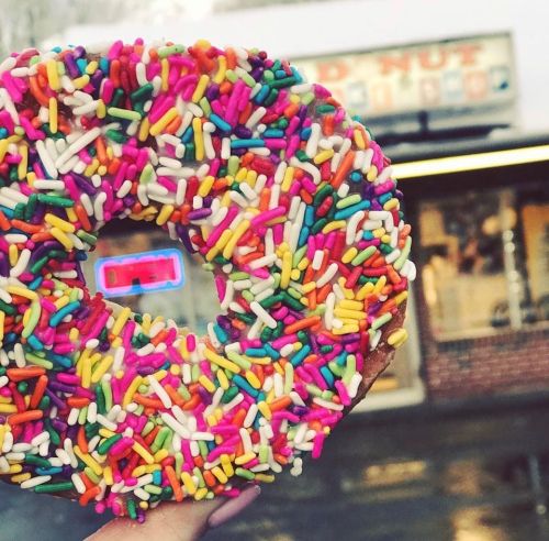 A large donut covered in colorful sprinkles, held up in front of a shop with an "OPEN" sign.