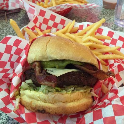 A close-up of a burger with bacon, lettuce, and pickles, served with a side of fries in a red-checkered basket.