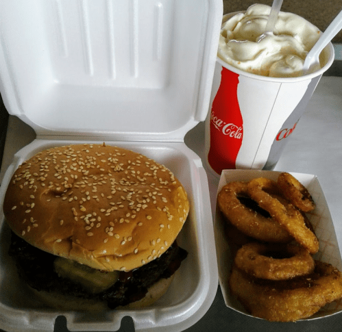 A sesame seed burger, onion rings, and a soft drink in a takeout container.