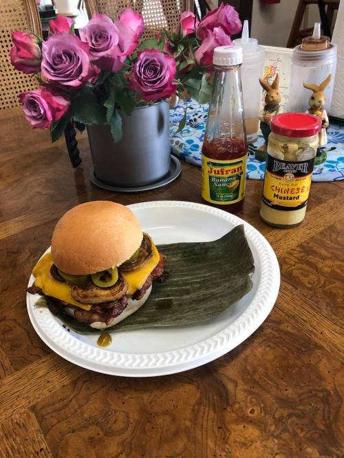 A cheeseburger with jalapeños on a plate, accompanied by mustard, hot sauce, and a vase of roses in the background.