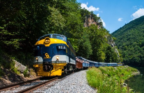 A vintage train travels along a scenic route, surrounded by lush greenery and mountains under a clear blue sky.