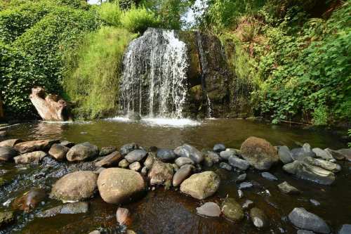 A serene waterfall cascades over rocks into a calm pool, surrounded by lush greenery and smooth stones.