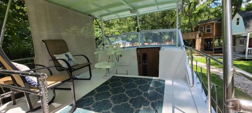 A cozy outdoor seating area on a boat, featuring chairs, a table, and a patterned rug, surrounded by greenery.