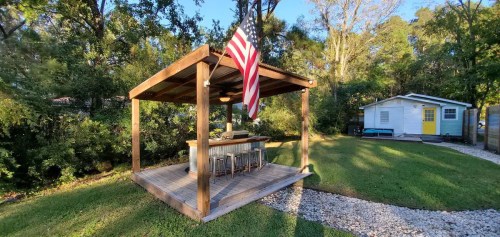 A wooden gazebo with an American flag, surrounded by greenery, next to a small blue house with a yellow door.