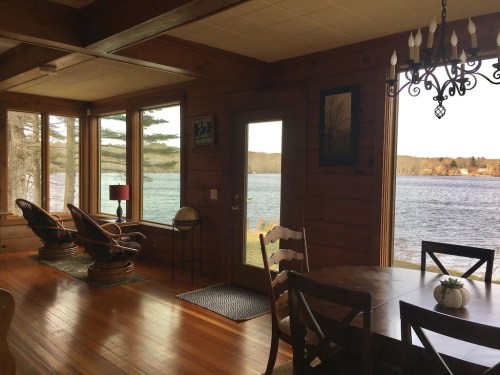 Cozy interior of a cabin with wooden walls, a dining table, and a view of a lake through large windows.