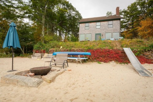 A sandy beach with a fire pit, chairs, a slide, and a house on a hill surrounded by trees and autumn foliage.