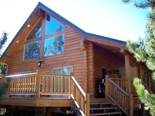 A log cabin with large windows and a wooden deck, surrounded by trees and blue sky.