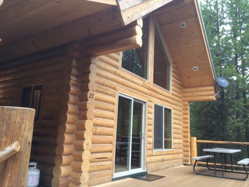 A wooden log cabin with large windows and a deck, surrounded by trees. A picnic table is visible on the deck.