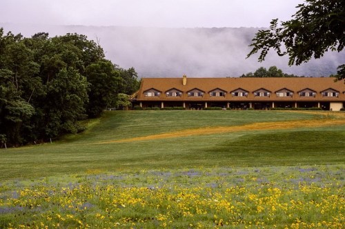 A scenic view of a lodge on a grassy field, surrounded by trees and misty mountains in the background.