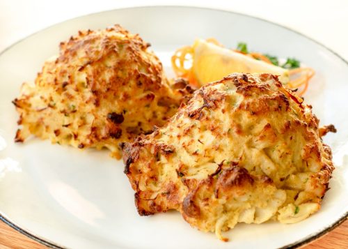 Two golden-brown crab cakes on a white plate, garnished with a lemon wedge and a sprig of parsley.