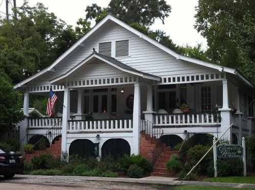 A charming white house with a large porch, American flag, and lush greenery surrounding it.