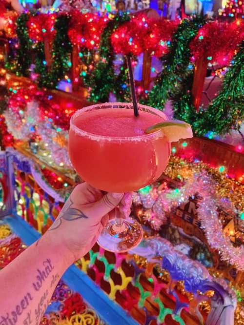 A vibrant cocktail in a large glass, garnished with a lime, held against a colorful, festive background of lights and decorations.