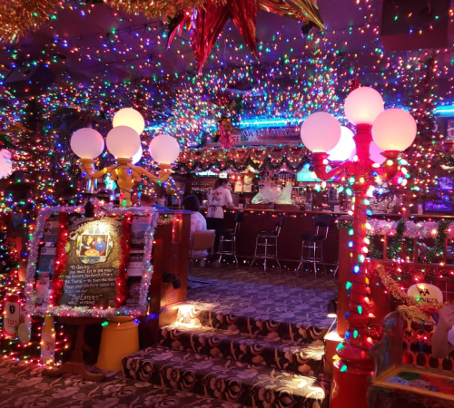A vibrant bar decorated with colorful Christmas lights, featuring a staircase and festive ornaments throughout.
