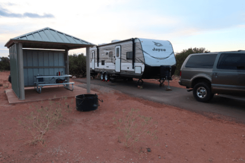 A camper trailer parked next to a shelter with a picnic table, surrounded by red dirt and sparse vegetation.