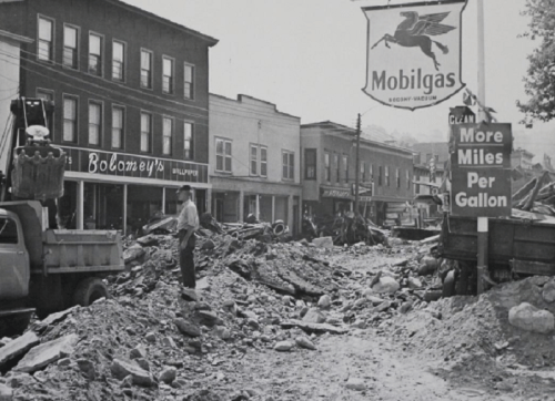 Historic black-and-white photo of a street covered in debris, with damaged buildings and a Mobilgas sign in the background.