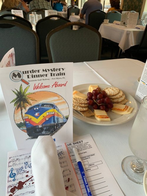 A table set for dining with a "Murder Mystery Dinner Train" menu, crackers, cheese, and grapes in the foreground.