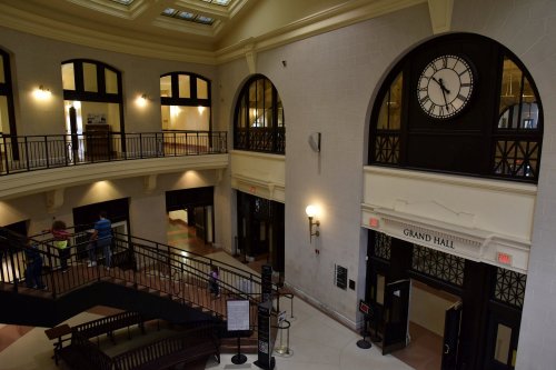Interior view of a spacious hall with large windows, a clock, and a staircase, featuring a welcoming atmosphere.
