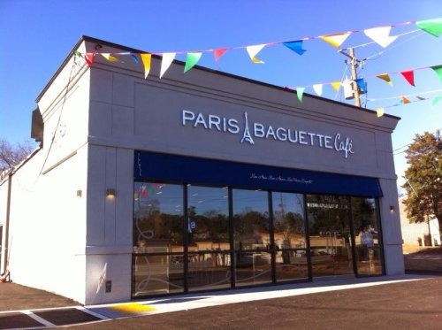Exterior of a Paris Baguette café with colorful flags and a clear blue sky.