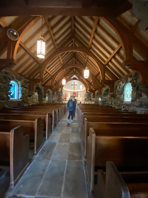 A person walks down the aisle of a rustic church with wooden beams and stone walls, surrounded by wooden pews.