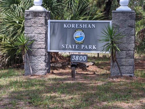 Sign for Koreshan State Park, featuring the park's logo and surrounded by greenery. Address: 3800.